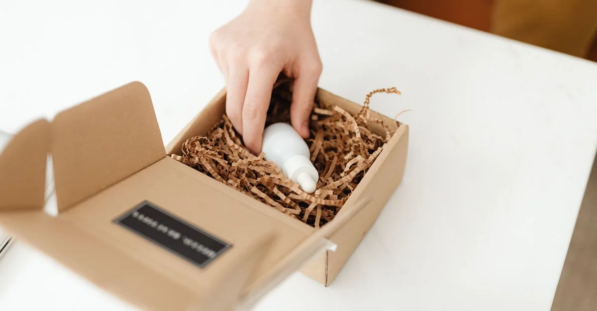 Gros plan d'une main déballant une boîte en carton avec une bouteille de produit à l'intérieur sur une table blanche.